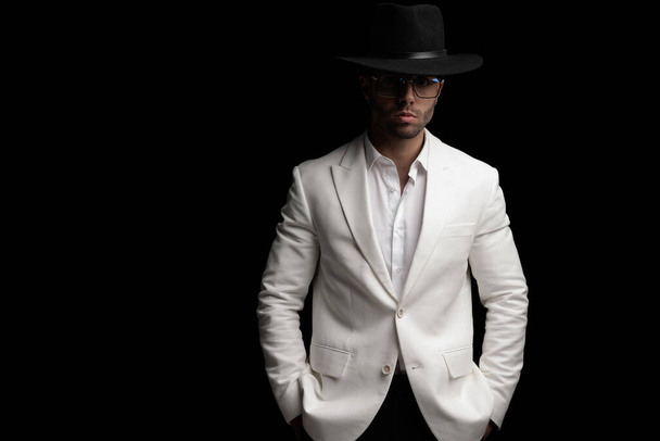 confident elegant man with hat and glasses holding hands in pocket and posing in front of black background - Φωτογραφία, εικόνα