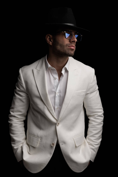 σέξι άντρας της μόδας σε λευκό σακάκι κοστούμι με ανοιχτό γιακά πουκάμισο φορώντας καπέλο και γυαλιά κοιτάζοντας προς τα πλάγια, ενώ θέτουν με τα χέρια σε τσέπες σε μαύρο φόντο - Φωτογραφία, εικόνα
