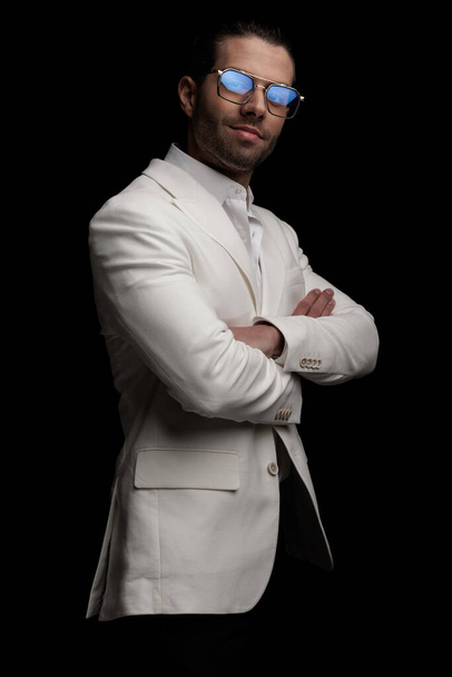 proud young man with glasses in white suit smiling and crossing arms and posing on black background - Foto, Imagen