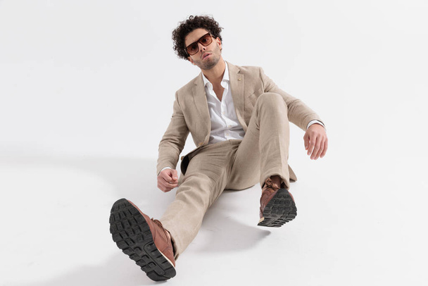 homem elegante atraente com camisa de colarinho aberto e terno bege sentado no chão em fundo cinza - Foto, Imagem