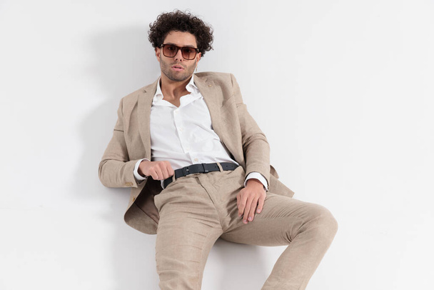 sexy uomo di moda con camicia collo aperto sdraiato a terra e in posa fiduciosa durante la regolazione cintura su sfondo grigio - Foto, immagini