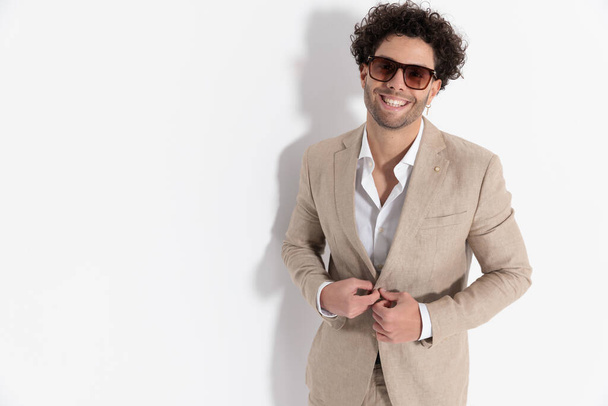 portrait of happy smart casual man with open collar shirt laughing and unbuttoning beige suit in front of grey background in studio - Fotó, kép