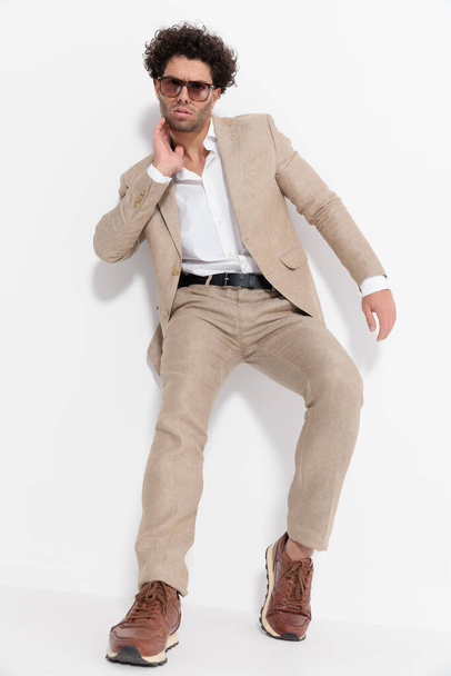 koele jongeman in beige pak met open kraag hand in hand op nek en poseren in een mode-manier op grijze achtergrond - Foto, afbeelding