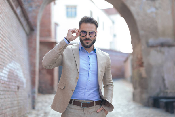 Egy fiatal üzletember portréja, amint a szemüvegét rendezgeti és kéz a kézben a zsebében tartja a szabadban, egy régi középkori városban. - Fotó, kép