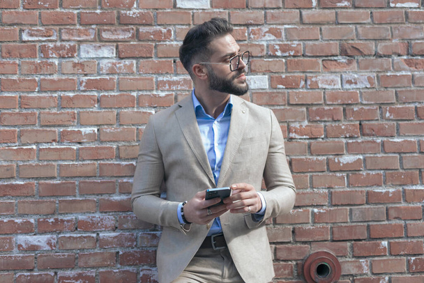 Portrait of young businessman texting on phone while leaning on brick wall and wearing eyeglasses outdoor, in an old medieval town - Fotó, kép