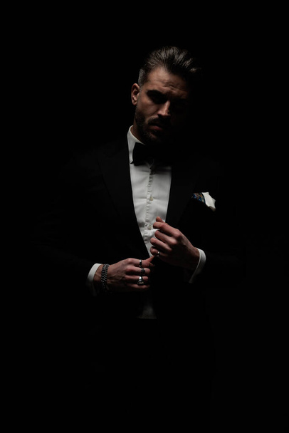 elegante elegante hombre ajustando y desabotonando esmoquin negro en frente de fondo negro - Foto, imagen