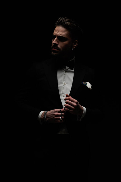 sexy bearded groom looking to side and adjusting black tuxedo in front of black background in studio - Fotó, kép