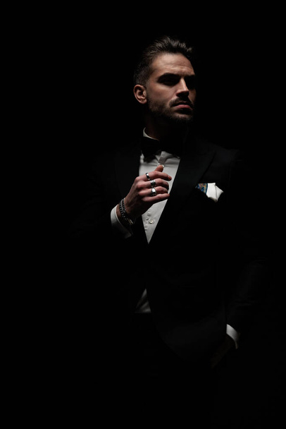 elegant bearded groom wearing black tuxedo and looking to side while posing with hand in pocket in a fashion way on black background - Photo, Image