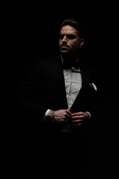 fashion young businessman looking to side and buttoning tuxedo while confidently posing on black background - Photo, Image