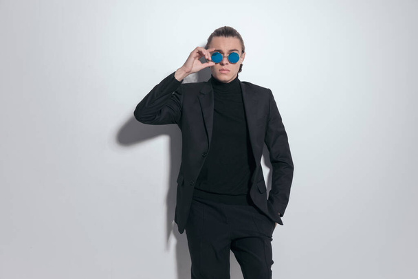 Imagem de moda de empresário atraente fixando óculos de sol com uma mão no bolso e vestindo uma roupa agradável contra fundo estúdio cinza - Foto, Imagem