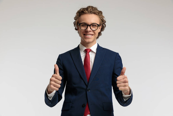 Портрет красивого бизнесмена, показывающего большой палец и одетого в официальный костюм на сером фоне студии - Фото, изображение
