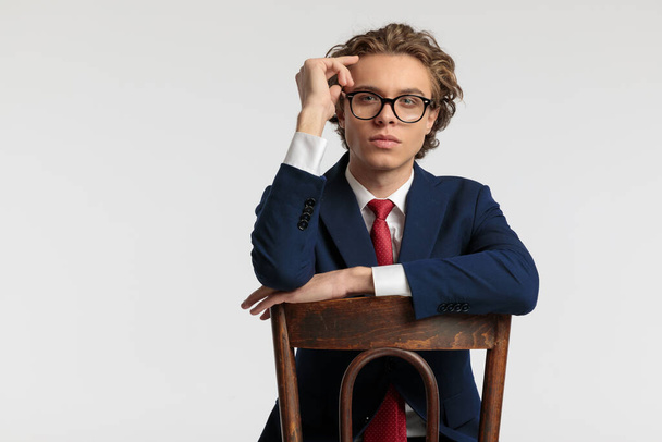 Portrait of sexy businessman leaning on wooden chair and wearing eyeglasses and wearing a formal outfit against gray studio background - Foto, imagen