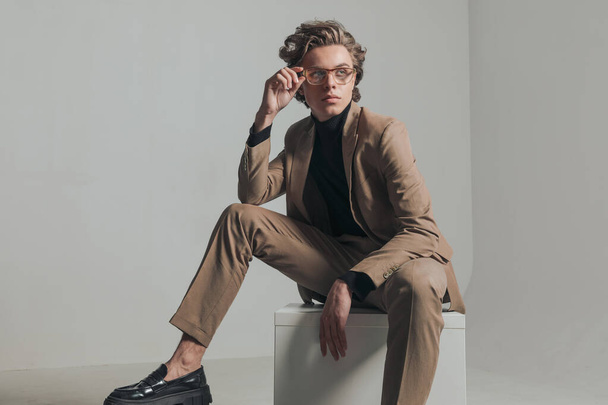 moda jovem com cabelo loiro olhando para o lado e ajustando óculos na frente de fundo cinza enquanto sentado com o joelho para cima - Foto, Imagem