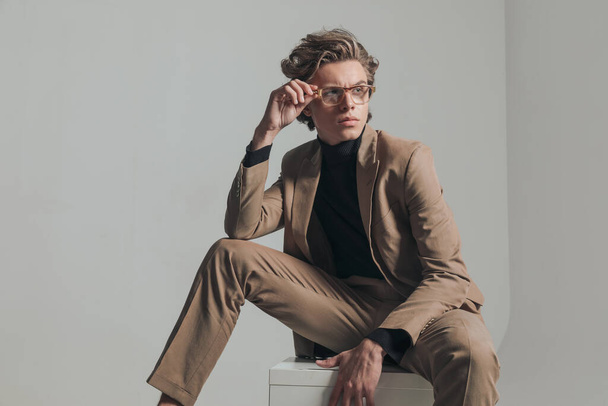 Smart Casual Man sitzt und hält den Ellbogen auf dem Knie, während er die Brille fixiert und auf grauem Hintergrund im Studio zur Seite schaut - Foto, Bild