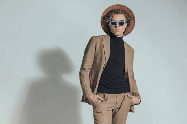 sexy fashion guy with hat and sunglasses holding hands in pockets in front of grey background in studio - Фото, изображение