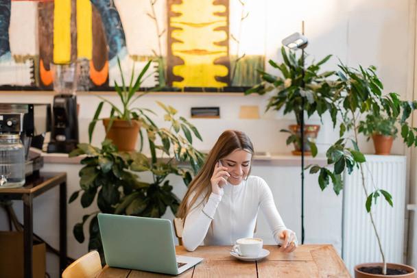 χαρούμενη blogger μιλώντας στο smartphone κοντά στο laptop δίπλα στο φλιτζάνι καπουτσίνο στο cafe  - Φωτογραφία, εικόνα
