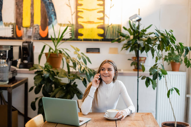 χαρούμενος ελεύθερος επαγγελματίας μιλώντας στο smartphone κοντά στο laptop δίπλα στο φλιτζάνι καπουτσίνο στο καφέ  - Φωτογραφία, εικόνα