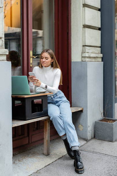 cheerful woman using smartphone near laptop and cup of coffee while sitting in cafe in Vienna  - 写真・画像