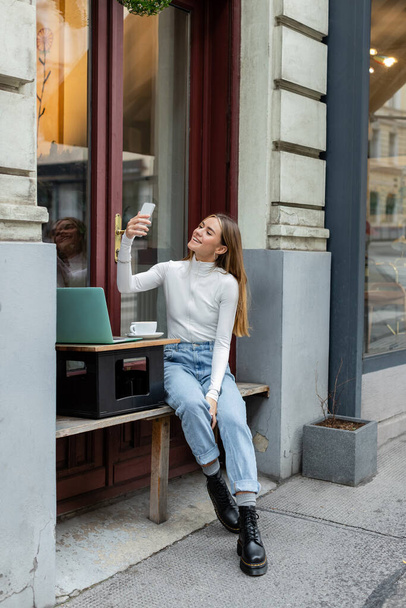 cheerful woman taking selfie near laptop and cup of coffee while sitting in cafe on street in Vienna  - Photo, Image
