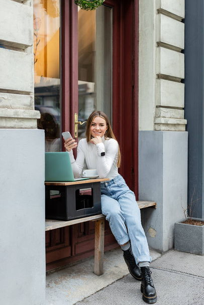 happy young woman holding smartphone near laptop and cup of coffee while sitting in outdoor cafe on street in Vienna  - Foto, Imagem