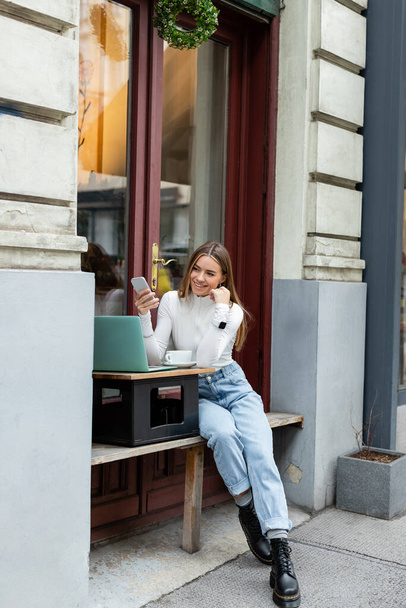 boldog nő kezében okostelefon közelében laptop és egy csésze kávé, miközben ül a kávézó az utcán Bécsben  - Fotó, kép