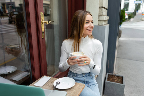 Бездоганна жінка, що тримає чашку капучино під час сидіння біля гаджетів на столі у кафе на вулиці у Відні.  - Фото, зображення
