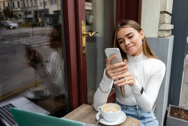 ευχαριστημένη γυναίκα κρατώντας smartphone κοντά σε φλιτζάνι latte στο τραπέζι, ενώ κάθεται σε υπαίθριο καφέ στη Βιέννη  - Φωτογραφία, εικόνα