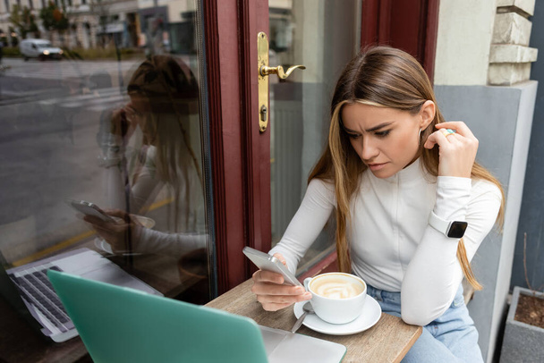 focused freelancer using smartphone near cup with cappuccino while sitting at bistro table near laptop in Vienna  - Φωτογραφία, εικόνα