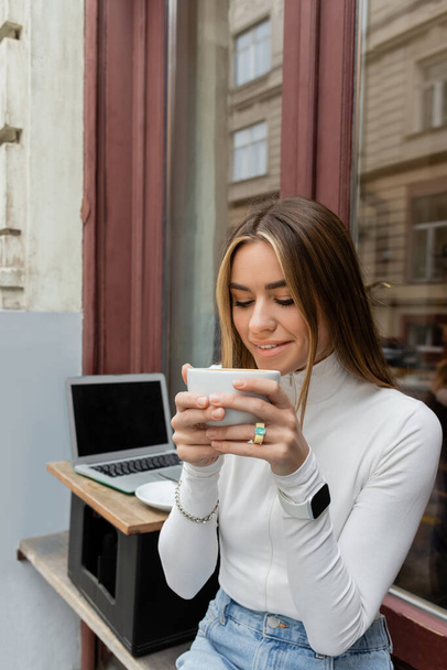 smiling woman holding cup of coffee while sitting in outdoor cafe next to laptop with blank screen in Vienna  - Photo, image