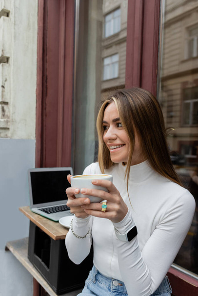 красивая женщина держит чашку кофе, сидя в кафе на открытом воздухе рядом с ноутбуком с чистым экраном в Вене  - Фото, изображение
