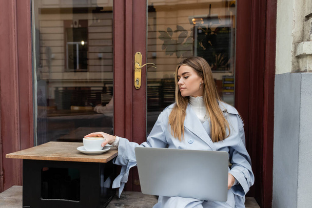 young freelancer in blue trench coat using laptop while reaching cup of coffee in outdoor cafe in Vienna   - Фото, изображение