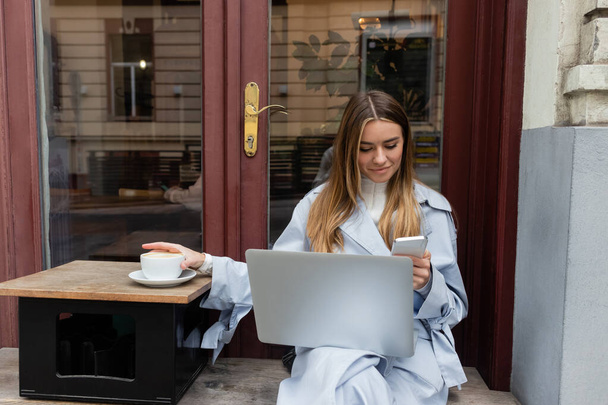 young woman in blue trench coat using smartphone near laptop while sitting next to cup of coffee in outdoor cafe in Vienna   - Φωτογραφία, εικόνα