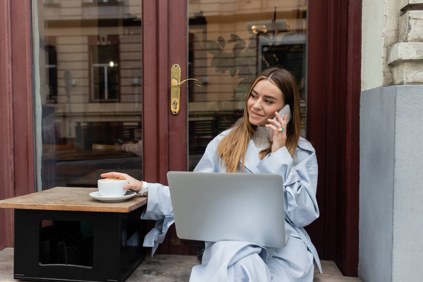 donna allegra in trench blu che parla su smartphone e siede accanto a una tazza di caffè in un caffè all'aperto a Vienna   - Foto, immagini