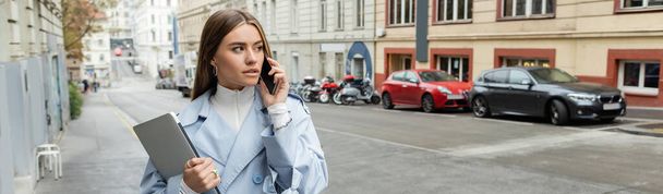 молодая женщина в синем пальто разговаривает на смартфоне, держа ноутбук на улице в Вене, баннер  - Фото, изображение