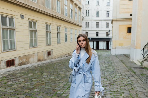 young woman in blue trench coat talking on smartphone while walking on street in Vienna  - Foto, imagen