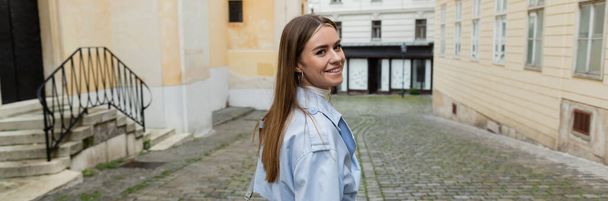 cheerful young woman in blue trench coat walking near buildings on street in Vienna, banner  - Foto, afbeelding