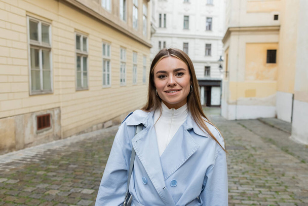 cheerful young woman in blue trench coat standing near buildings on street in Vienna  - Foto, Bild