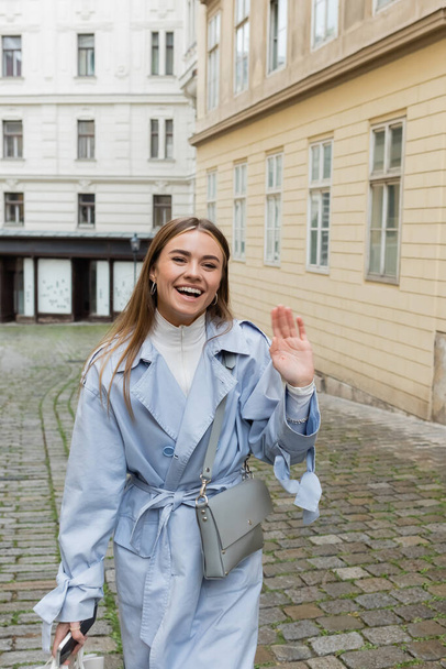 mujer feliz en gabardina azul agitando la mano mientras camina con el teléfono inteligente cerca de los edificios en Viena  - Foto, Imagen