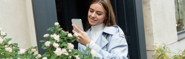 happy young woman in blue trench coat taking photo of green bush with blooming flowers on street in Vienna, banner  - Photo, Image