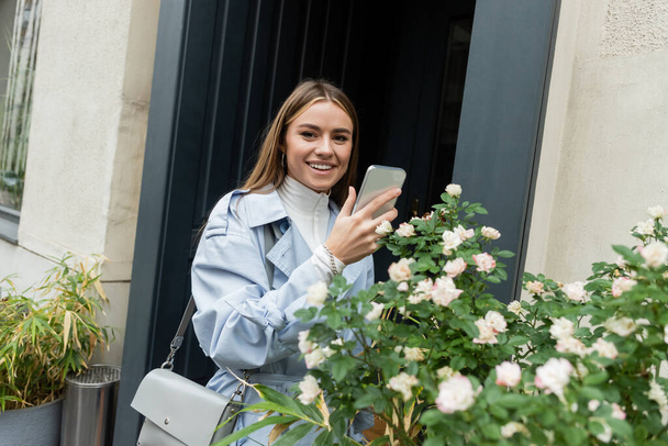 alegre joven en gabardina azul tomando fotos de arbusto verde con flores florecientes en la calle en Viena  - Foto, Imagen