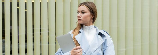pretty woman in blue trench coat holding laptop while standing near window shades on street in Vienna, banner  - Photo, Image