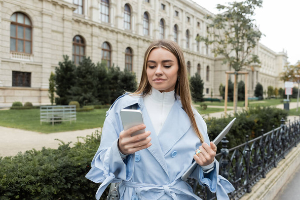 young woman in blue trench coat holding laptop and smartphone near historical building in Vienna  - Fotó, kép