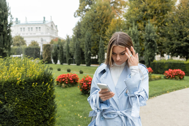 young woman in blue trench coat using mobile phone while standing in green park in Vienna  - Foto, Imagem