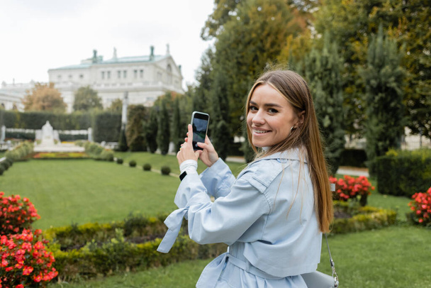 スマートフォンで緑の公園の写真を撮るブルートレンチコートの幸せな若い女性  - 写真・画像