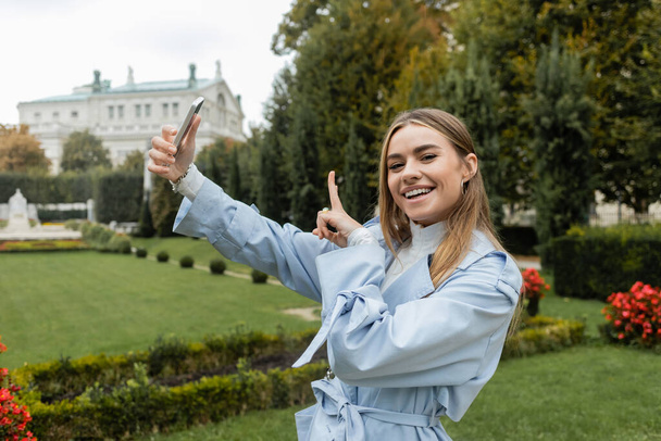 happy young woman in blue trench coat pointing away while using smartphone in green park in Vienna  - Φωτογραφία, εικόνα