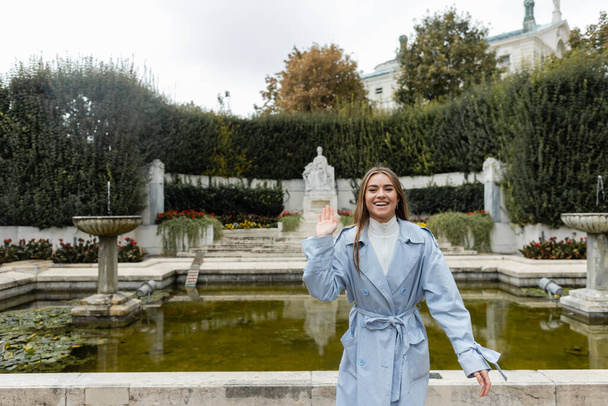 smiling young woman in blue trench coat waving hand near fountain in green park  - 写真・画像