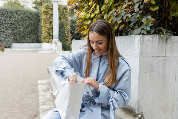 elégedett nő kék ballonkabátban ül a betonpadon, és belenéz a bevásárló táskába a bécsi zöld parkban  - Fotó, kép