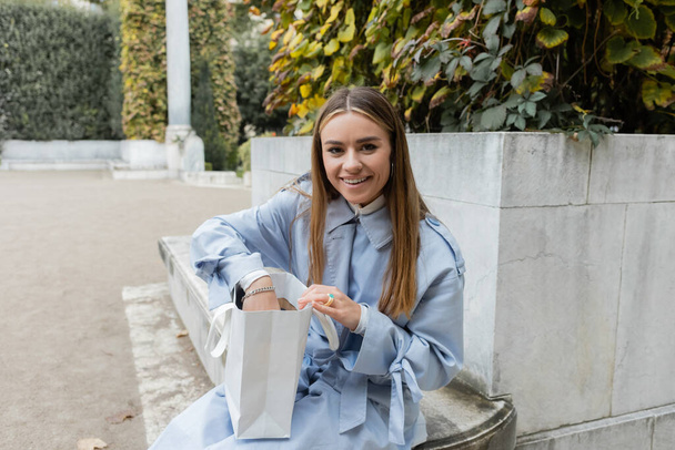 šťastná mladá žena v modrém plášti sedí s papírovým sáčkem na betonové lavičce v zeleném parku ve Vídni  - Fotografie, Obrázek