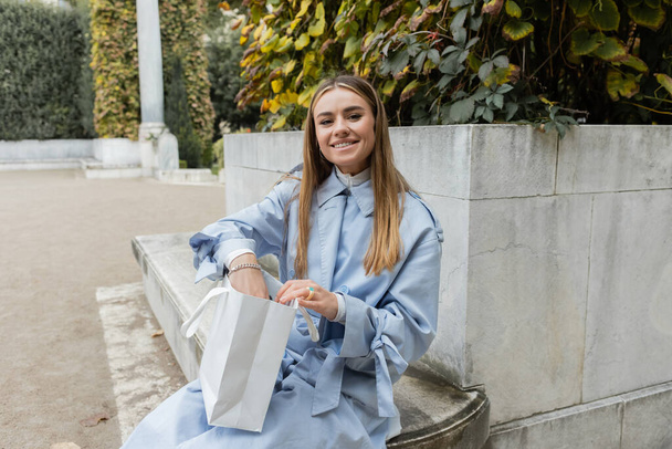 smiling young woman in blue trench coat sitting with shopping bag on concrete bench in green park in Vienna  - Photo, Image