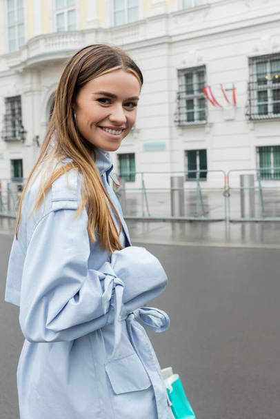 iloinen nuori nainen sininen trenssi takki hymyilee kaupunkien kadulla Wienissä  - Valokuva, kuva